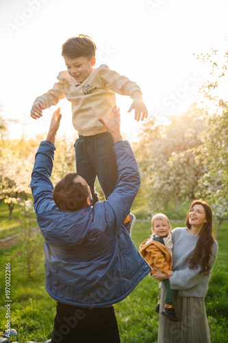 Happy family of four in a sunny spring garden. Father throwing eldest son in the air, mother holding youngest toddler son and watching. Active leisure, parenthood, childhood and love moments.