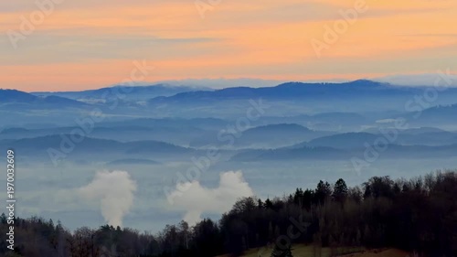 Layered blue mountain range shrouded in morning fog under a soft orange sky, with industrial smoke plumes rising from the valley, representing environmental impact