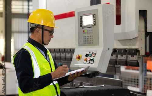 An Asian man, an industrial engineer by profession, wearing a safety helmet, operates machinery and records inspection logs in a factory.