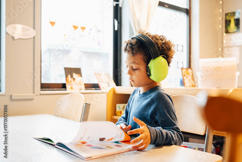 Boy wearing green ear muffs while reading book at table in kindergarten classroom