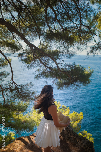 Back view of girl in summer outfit with flowing skirt and hat twirling skirt on cliff with Adriatic waves crashing and green Montenegro hills background