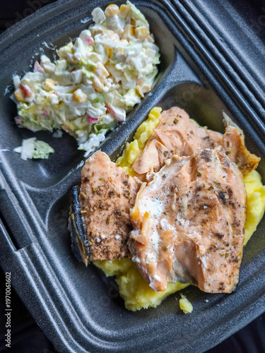 Cooked pink salmon pieces seasoned with herbs rest atop yellow mashed potatoes beside creamy vegetable salad containing corn and crab sticks inside black disposable food container.