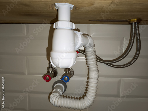 Close up view of white plastic drain siphon with flexible corrugated pipe connected to bathroom plumbing against white subway tile wall background under wooden vanity surface.