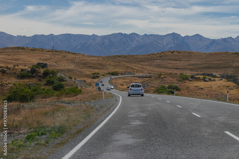 Fototapeta premium Winding Road Through Arid Hills with Distant Mountains Under Cloudy Sky