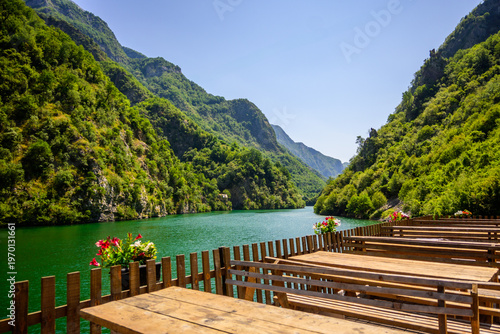 A charming outdoor restaurant terrace with rustic wooden tables and benches, offering expansive views of the vibrant green Koman Lake and forested mountains in Albania