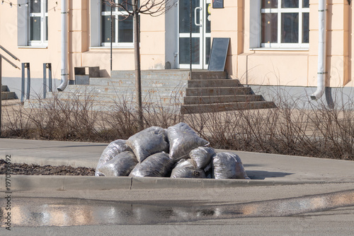 Pile of grey plastic garbage bags left on a city sidewalk next to a puddle with reflection.Spring city cleanup results: sacks filled with waste and dry leaves waiting for disposal.