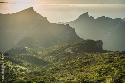 Mountain Landscape Tenerife With Winding Road, Dramatic Cliffs, Sun Rays and Green Hills in Misty Evening Light, Scenic Nature View of Rugged Terrain and Deep Valleys