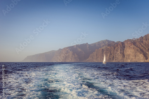 Atlantic ocean seascape with sailboat near dramatic volcanic cliffs of Tenerife island, Canary Islands coastline, blue water wake and horizon, travel adventure.