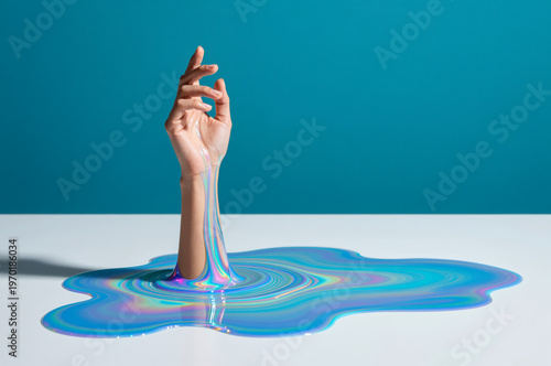 Human hand emerges vertically from viscous iridescent fluid on white surface against teal backdrop