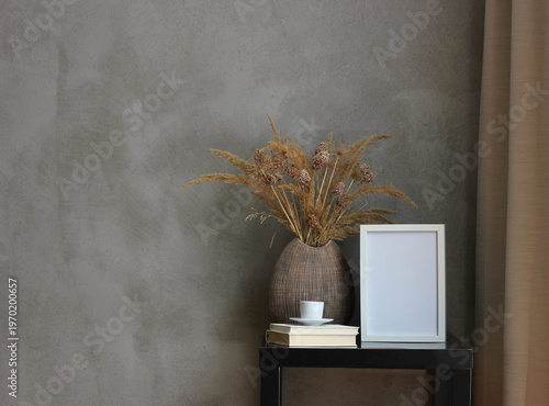 Minimalist interior mockup with empty white frame on small table, ceramic vase with dried grasses, books and coffee cup against dark gray wall, modern Scandinavian home decor scene with copy space