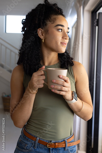 Woman standing near sliding glass door, holding ribbed ceramic mug and gazing at sunlight