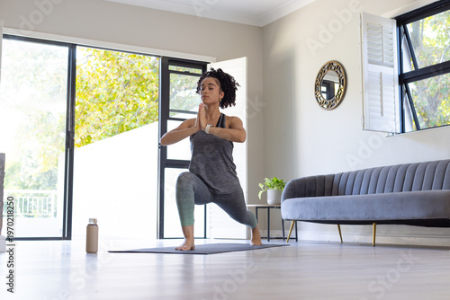 Woman practicing low lunge on yoga mat in living room wearing sportswear near water bottle