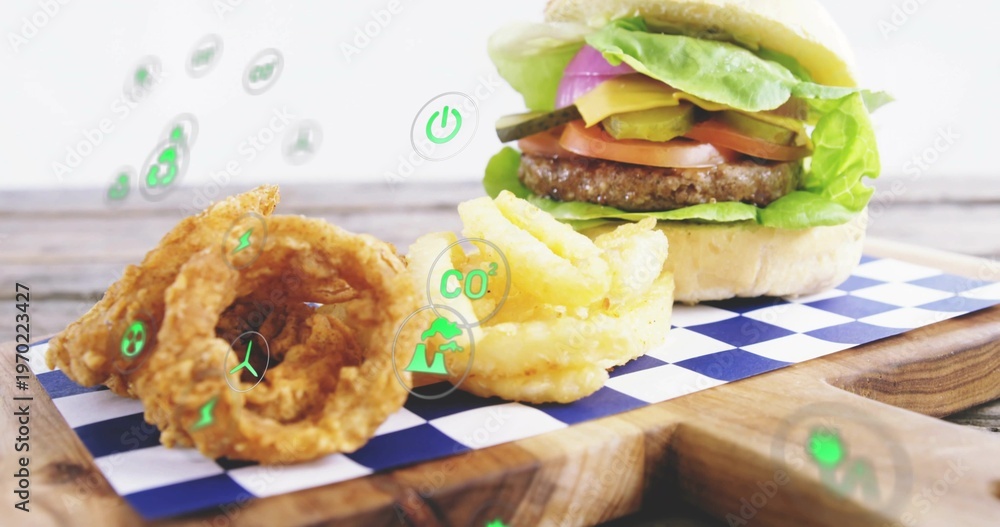 Fototapeta premium Displaying cheeseburger on board in studio, checkered paper, onion rings, fries, floating eco icons