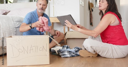 Senior couple on bedroom floor using tablet, sorting clothes for donation into DONATION box