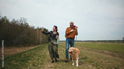 A couple walks on a path while their golden retriever enjoys the outdoor activity. They hold drinks and take photos in a natural setting.