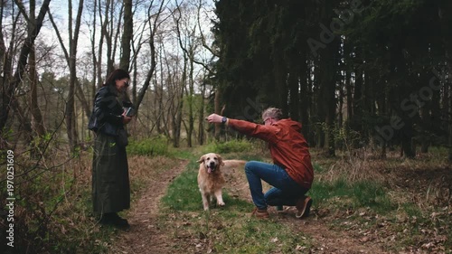 Two people stand on a dirt path while a golden retriever runs towards them in a forest setting during daytime.