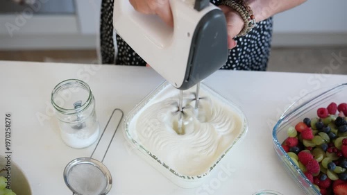 Girl is using a hand mixer to blend cream in a bowl while preparing a fruit dessert in a kitchen.