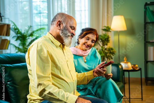 Canvas Print Retired Asian couple sitting on a sofa while sharing a smartphone for digital tasks at home