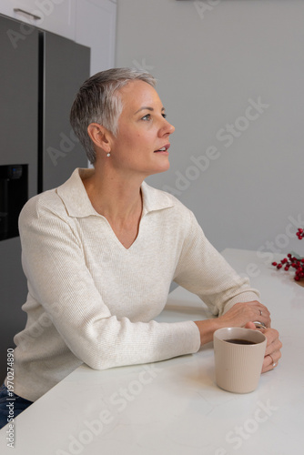 Senior woman sitting at kitchen counter holding beige mug on countertop in cream sweater, looking