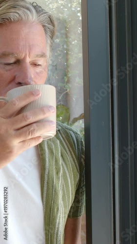 Vertical video: Checking aroma of ribbed mug, man sipping at home by glass door in striped shirt