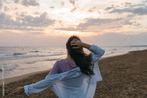Fotografi Young woman stands in profile on beach, holding hands to head as the wind blows her long hair