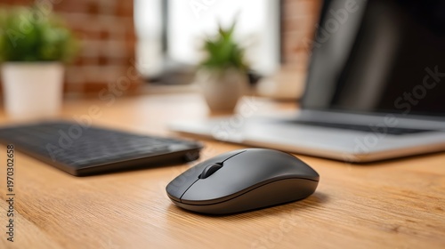 A modern wireless computer mouse rests on a wooden office desk beside a keyboard and laptop