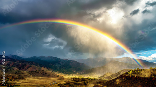 Rainbow arcing over mountains with a rain shower in a valley. Golden sunlight breaking through dark clouds