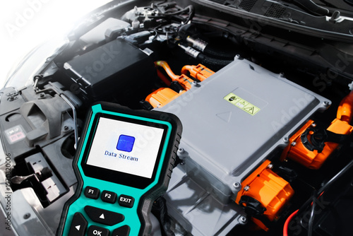 Electric Car Diagnostic Scanner, A teal diagnostic scanner shows Data Stream while held over a modern electric car engine with orange high-voltage cables in a workshop.