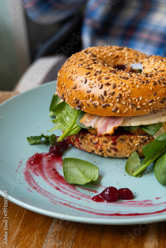 Delicious bagel sandwich with bacon, fried egg, baby spinach and arugula with cranberry jam on a blue plate in a cafe