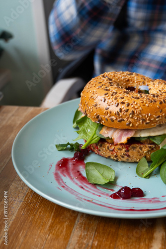 Delicious bagel sandwich with bacon, fried egg, baby spinach and arugula with cranberry jam on a blue plate in a cafe