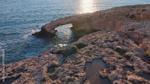 Aerial drone view of the Bridge of Love, a natural rock arch formation in Ayia Napa, Cyprus. Turquoise waves flow beneath the limestone arch