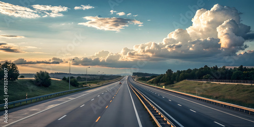 Straight multi lane motorway extends toward horizon through green rolling hills beneath expansive blue sky filled with giant fluffy white clouds during soft daylight.