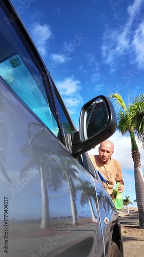 Young bald man washing and wiping the side window of his modern car parked on a road with palm trees, using a spray cleaner and a microfiber cloth on a sunny day with a blue sky