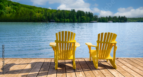 Rear view of two bright yellow Adirondack chairs on a weathered dock in Muskoka Ontario Canada, calm blue lake and forested hills, cottage country, no people, copy space.