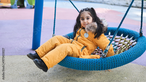 Young girl in winter clothes happily swinging on a nest swing at a colorful playground