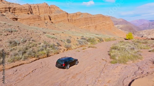 Black station wagon navigates a dirt road through a serene desert landscape. Eroded rock formations and distant mountains create an adventurous atmosphere. Following drone view.