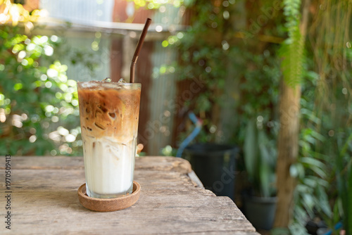 Glass of iced latte with milk and espresso layers served on a wooden table in a garden setting with lush green background and soft sunlight.
