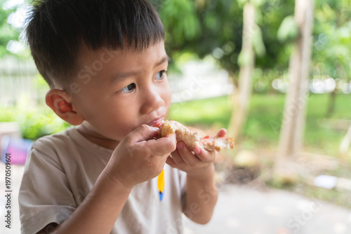 A young Thai boy eating a slice of bread with jam outdoors in a natural setting.