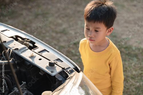 A thoughtful child in a yellow shirt observes a car engine under the hood, learning about mechanics outdoors.