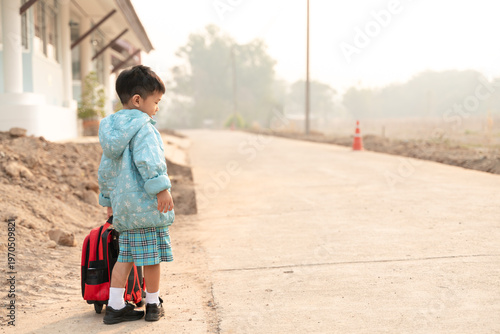 Young Thai child in a school uniform and winter coat standing on a rural road with a backpack in the morning light.