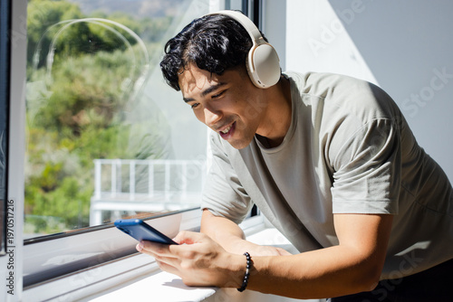 Adult man leaning on windowsill at home smiling checking blue phone with headset and bracelet