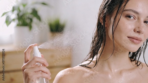 Young woman with wet hair sprays facial mist from white bottle onto skin in sunlit bathroom with wooden shelf and green potted plants nearby