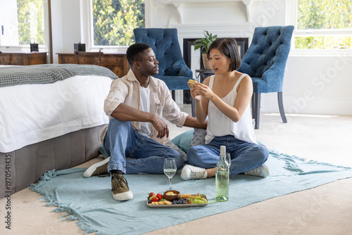 Diverse couple sitting on light-blue blanket in bright bedroom, sharing smartphone and sipping wine