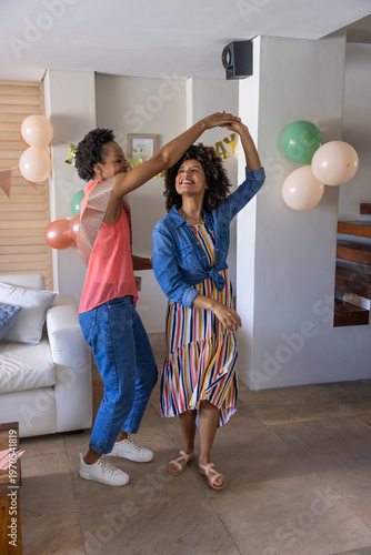 Diverse female friends dancing together in living room near balloons, banner, white sofa