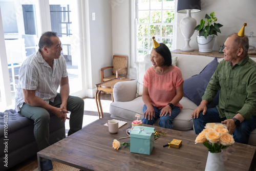 Asian family with seniors celebrating birthday at home at coffee table with lit cupcake, party hats