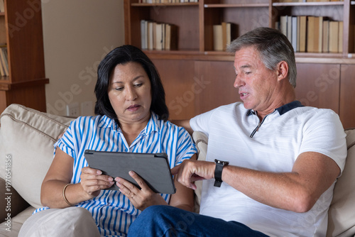 Diverse couple sitting on beige sofa in living room, holding black tablet and pointing at bookshelf