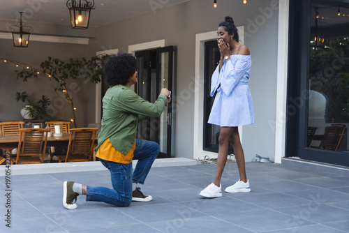 African American couple kneeling on patio, proposer holding open ring box, string lights glowing