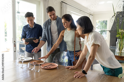 Diverse group with tablet pointing and arranging pink plates at dining table in modern home