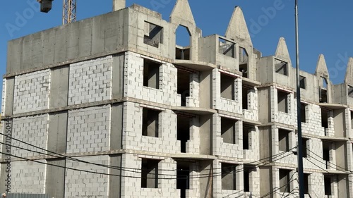 Unfinished apartment building with aerated concrete blocks under blue sky, exposed balconies and repetitive window openings, crane.