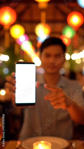 A person holds a blank screen smartphone in a dimly lit restaurant with colorful lanterns in the background